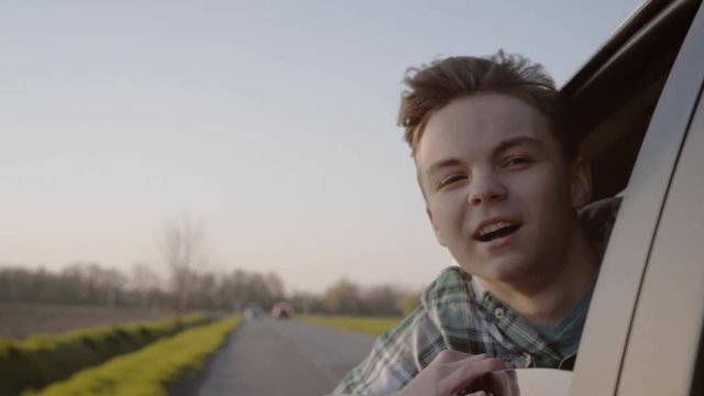 Carefree Teen boy looking out car window and smiling. Happy child enjoying leisure in automobile during sunny day road trip. Slow motion.