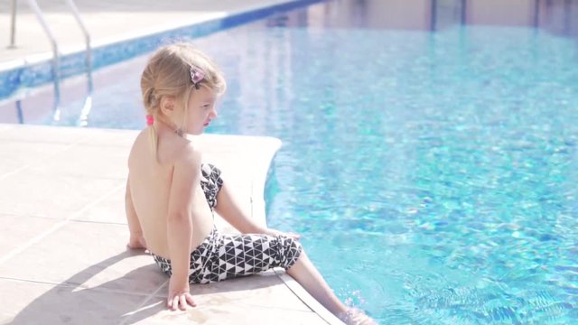 Beautiful little girl ploping feet in the pool. She sits by the swimming pool and sprinkles her legs.
