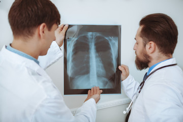 Obraz premium Rear view shot of a two male doctors examining x-ray scan of a patient. Medical team discussing lungs x-ray of a patient. Experienced therapists working at the clinic. Medical service concept