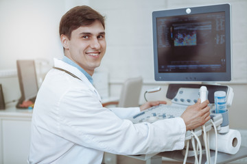 Fototapeta na wymiar Happy handsome male doctor smiling to the camera, working on ultrasound scanning machine at his clinic. Experienced doctor working at modern medical facility with high quality equipment