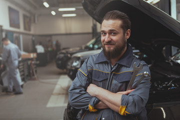 Cheerful bearded mechanic in grey uniform smiling to the camera, holding a wrench. Friendly car repairman resting at thegarage after examining broken car, copy space