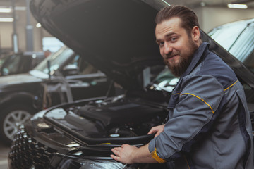 Obraz premium Cheerful bearded mechanic smiling over his shoulder to the camera, while repairing an SUV car at the garage, copy space. Friendly auto technician enjoying working with automobiles