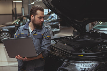 Experienced car technician using laptop, during car diagnostic at his workshop. Bearded male...