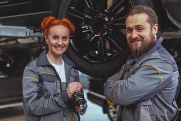 Fototapeta na wymiar Cheerful beautiful female mechanic and her male colleague smiling to the camera, working at their car service station. Experienced car technicians repairing vehicles at the garage. Female equality con