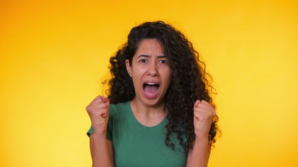 Frightened hispanic woman with curly hair in green t-shirt afraid of something and looks into the camera with big eyes full of horror over yellow background.