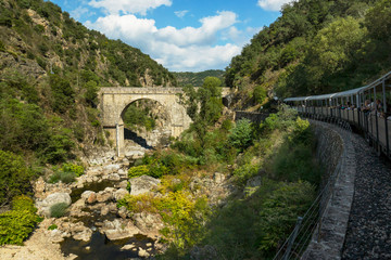 Fototapeta na wymiar Old railway line in french Ardeche region with tourist wooden train in a foreground and the massive stone bridge in a background. France 2018.