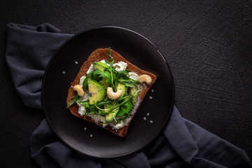 Toasted toast with ricotta cheese, avocado slices, arugula, spinach and cashew nuts on a black...