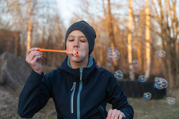 a teenage boy in a blue jacket and a gray hat is blowing bubbles in the open air. children's portrait