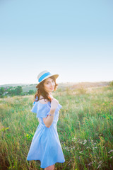 Fototapeta na wymiar Portrait of young woman in blue romantic dress and straw hat on green meadow enjoying summer sunset. Calm and harmony. Summer vacation,fun, positive mood. Back sun light, sun beams. Copy space.