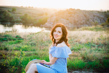 Portrait of young woman in blue romantic dress and straw hat in sun light enjoying summer sunset on nature. Calm and harmony. Summer vacation,fun, positive mood. Back sun light, sun beams. Copy space.