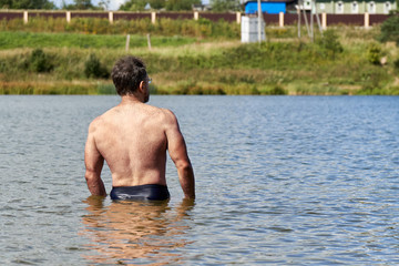 Sportive middle-aged man is bathing in the small suburban lake in summer. Back view