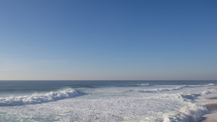 Empty long wide beach with yellow sand in Nazare; big portugal curling wave crashing into sandy shore; crystal clear drops of seawater fly in the air; empty emerald tube wave; nature scenic beauty