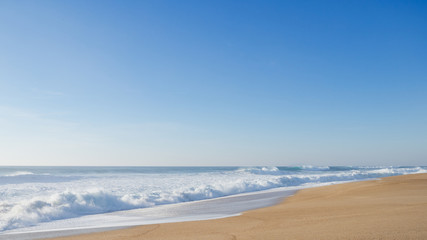 Empty wide coastline with yellow sand in Nazare; huge curving waves crushing on the beach; sunny evening with cloudless sky; clear skyline; sea foam all the way out to the horizon, Portugal