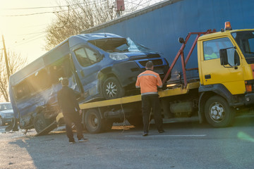 Loading a broken blue minibus on a yellow tow truck in the middle of the street on a sunny day