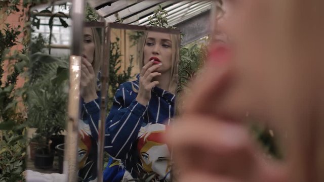 Beautiful blue-eyed blonde admires herself in mirror. She is in botanical garden greenhouse wear striped blue jacket, touching your fingers to your lips. In the background is pink wall and green trees