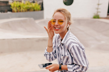 Hippie happy girl with blond hair wearing orange glasses listening music in the headphones and holding smartphone 