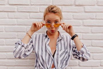 Funny adorable lady with blond hair posing to camera while making faces and holding glasses while sending a kiss