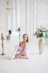 A cute  ballerina in a pink ballet costume is sitting on the floor and tying pointe shoes. Ballerina in the dance class.The girl is studying ballet. Ballerina dancing in a white studio. ballet tutu