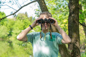Horizontal color photography of cute white funny little kid looks through old vintage black binoculars at camera smiling  while standing at scenic blurry green fresh foliage of spring trees background