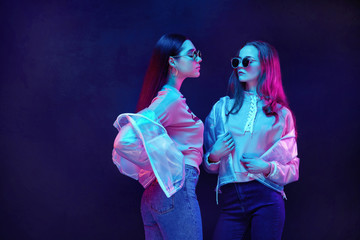 Contemporary teen girls wearing sunglasses with plastic coats posing in neon lights