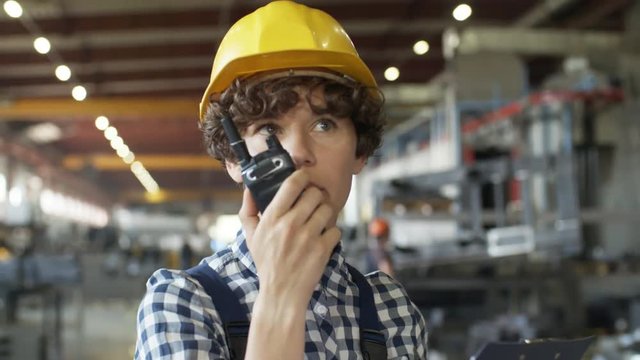 Pan chest-up shot of curly 30-something female factory engineer with clipboard, overseeing work and giving directions on portable radio, standing in spacious workshop with lots of machinery