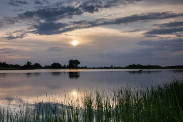 Sun in the clouds over the horizon. Calm lake and grass