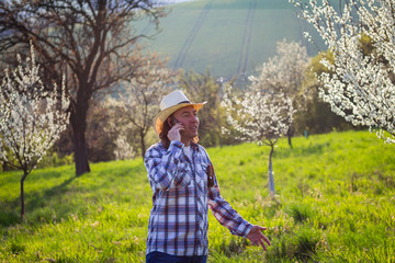 Smiling farmer talking to smart phone while examining blooming fruit trees in orchard