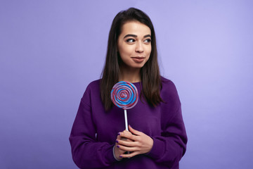 Portrait of happy young female holding a purple lollipop isolated over violet background