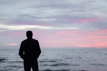 Young adult man in warm black overcoat standing alone and staring at small waves of sea and overcast sky. Peaceful atmosphere in winter evening. Back view. Empty place for text, quote or sayings.
