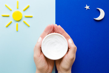 Young, perfect woman hands holding white jar of moisturizing cream. Care about nails and clean, soft, smooth body skin in 24 hours day. Sun and moon shapes on light and dark blue background. Closeup. 