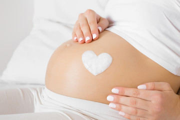 Girl sitting in bed. Young woman's hands touching her naked big belly. Heart shape created from...