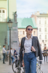 Woman in jacket and jeans walking on the street. Cheerful stylish woman with sunglasses outdoor. Woman wearing shades while looking away in the city street