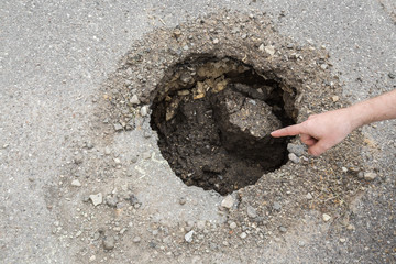 Driver's hand pointing to the big, deep hole on the asphalt road. Dangerous, bad automobile driveway. Damaged surface. 