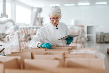 Young Caucasian employee in sterile uniform holding tablet and counting products in boxes. Food...
