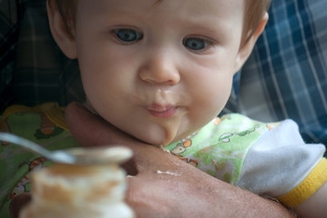 A little baby girl sits in the arms of her grandfather and eats a fruit puree with a spoon. Introduction of complementary foods in a child. Big rough hands gently support the baby
