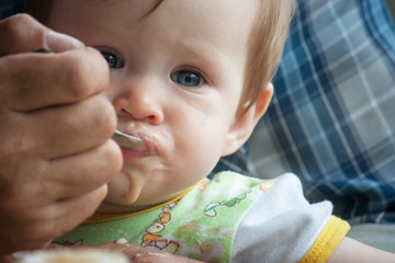 A little baby girl sits in the arms of her grandfather and eats a fruit puree with a spoon. Introduction of complementary foods in a child. Big rough hands gently support the baby