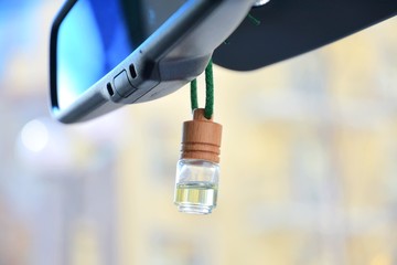 Car air perfume freshener bottle inside the car with part of car mirror on blurred city background. Little glass bottle with wooden lid and yellow aromatic liquid automobile freshener on a green rope.