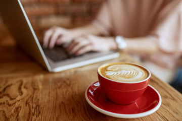 Close up of fresh espresso in red cup. In background woman typing on report on laptop.
