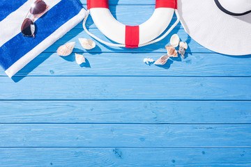 top view of striped towel, sunglasses, lifebuoy, floppy hat and seashells on blue wooden background with copy space