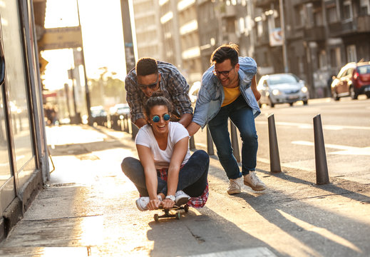 Group of friends hangout at the city street.Female sitting on skate board while friends pushing her from behind.