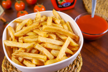 French fries in white bowl on a braided pad on wooden table with tomato and ketchup next to it