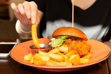 Woman eating burger with hand dipping french fried to ketchup.
