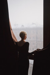 A beautiful slender young woman with light short hair in a black dress stands at the window in a room with dark curtains. Vertical picture.