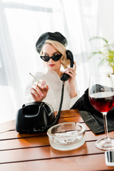 selective focus of elegant blonde woman in black beret and sunglasses smoking and talking retro phone near glass with red wine