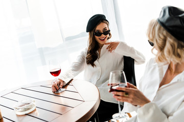 elegant smiling and surprised brunette woman in black beret and sunglasses drinking red wine at wooden table with blonde friend