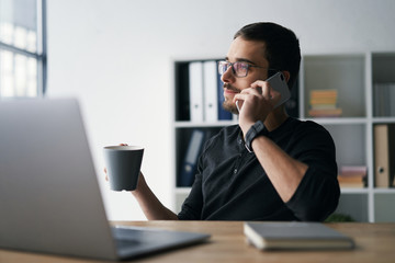 Young man working with phone and computer, receiving phone call, talking with partners 