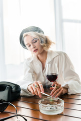 selective focus of elegant blonde woman in black beret smoking cigarette and holding glass with red wine