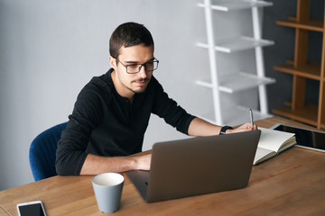 Young man working with computer, phone and tablet at the table while drinking coffee