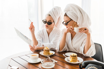stylish women in bathrobes, sunglasses and jewelry with towels on heads smoking cigarette and...