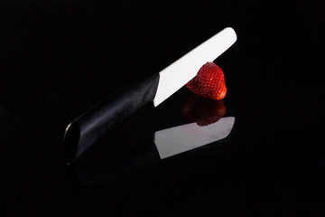 A white blade knife cuts a fresh strawberry and its reflection with black background.